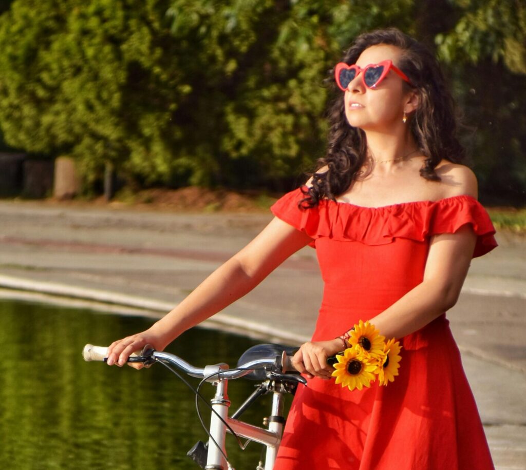 Stylish young woman in a red dress with sunflowers posing with a bicycle by a pond in a sunny park.