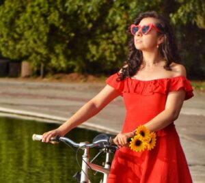 Stylish young woman in a red dress with sunflowers posing with a bicycle by a pond in a sunny park.